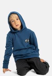 Modrá mikina s kapucňou z jemného materiálu, s čiernym logom na hrudi, elastickými manžetami a klokankou. Nosí sa s čiernymi nohavicami.
