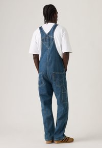 Ολόσωμη φόρμα τζιν σε μέτριο μπλε χρώμα, με ρυθμιζόμενες τιράντες, πολλαπλές τσέπες και άνετη εφαρμογή. Συνδυασμένη με άσπρη μπλούζα και καφέ παπούτσια.