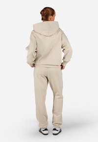 Μπεζ φούτερ με κουκούλα και συνδυασμένα φόρμα πάνες, από βαμβακερό μείγμα, άνετη εφαρμογή, ριμπ μανσέτες και κορδόνι στην κουκούλα. Μαύρα και άσπρα αθλητικά παπούτσια.