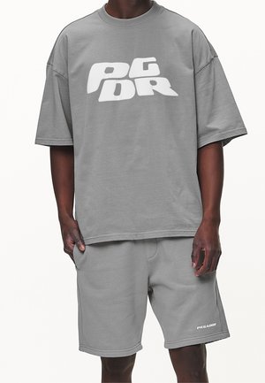 Man draagt een loszittend grijs T-shirt met witte blokletters en bijpassende grijze shorts met een klein wit logo op het linkerpantbeen.