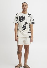 Camiseta de algodón color beige claro con un gran estampado floral negro, combinada con pantalones cortos color crema. Presentada sobre un fondo neutro con detalles mínimos.