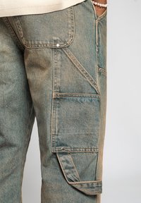 Denim cargo-byxor i en urtvättad blågrön nyans med flera fickor, kontrasterande sömmar och en texturerad yta. Utrustad med ett tryckknappsstängning.