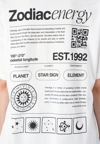 T-shirt branca com texto preto, apresentando símbolos astrológicos e um código QR, detalhando a energia zodiacal, planetas e motivos celestiais.