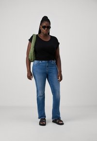 Black t-shirt, blue straight-leg jeans, black sandals, green quilted bag, and dark sunglasses. Smooth textures and casual fit.