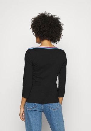 Persoon met krullend haar, gekleed in een zwart shirt met lange mouwen met blauwe en rode biezen bij de halslijn en een blauwe spijkerbroek, met de rug naar voren.