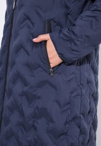 Cappotto imbottito blu scuro con texture trapuntata, caratterizzato da una tasca con zip e dettagli in zip neri sul davanti, design su misura e aderente.