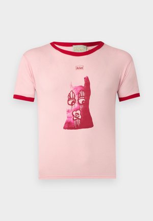 Młody mężczyzna z kręconymi włosami, ubrany w różowy obcisły T-shirt z czerwonym obszyciem i grafiką, wyblakłe jeansy z jasnymi nadrukami oraz brązowe buty z pomarańczowymi sznurówkami.
