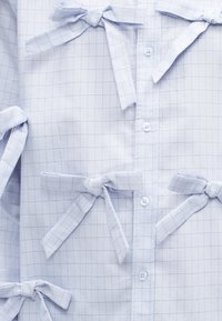 Camisa de padrão quadriculado azul claro com frente abotoada e detalhes amarrados. Feita de tecido suave com linhas quadriculadas mais escuras contrastantes.