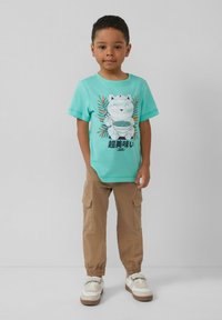 Turquoise katoenen t-shirt met een grafisch motief van een kat met noedels, gecombineerd met beige cargobroeken en witte sneakers met bruine zolen.