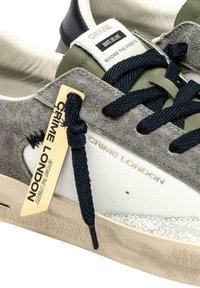 Ingrandimento di una sneaker Crime London grigia e bianca con lacci neri, texture in suede e pelle, e un'etichetta gialla con il marchio sul lato.