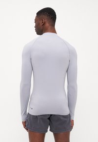 Világosszürke, hosszú ujjú rash guard, sima textúrával és testhezálló kialakítással, lapos varratokkal és kerek nyakkivágással, sötét rövidnadrággal párosítva.
