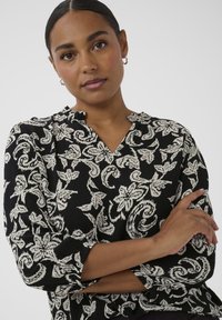 Frau mit dunklem, zurückgebundenem Haar, die silberne Creolen trägt und eine schwarze Bluse mit weißen Blumen- und Paisleymustern anhat, mit verschränkten Armen.