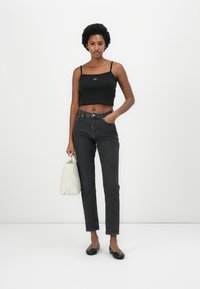 Black cropped tank top with a label, high-waisted dark denim jeans, and black flats. Holding a textured white handbag with quilted detailing.