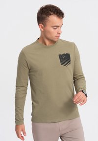 Olivgrünes Langarmhemd mit einer Brusttasche, das ein schwarzes Grafikdesign und horizontale Streifen zeigt. Weiches, glattes Material.