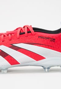 Chaussure de soccer rouge avec une tige texturée, arborant trois rayures blanches et des accents noirs. Comprend des lacets et une semelle transparente avec des crampons.