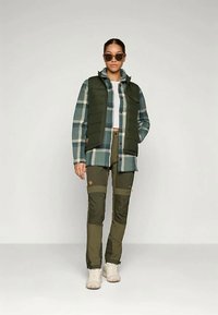 Vihreä tikkivesti sinivihreän ruudullisen paidan päällä, yhdistettynä oliivinvihreisiin taskuhousuihin, joissa on polvilaikkuja, ja valkoiset lenkkarit.