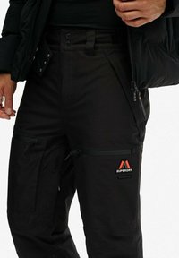 Nærbillede af en person, der har sorte Superdry Mountain-bukser på med lynlås-lommer og en sort jakke, der er delvist åbnet.