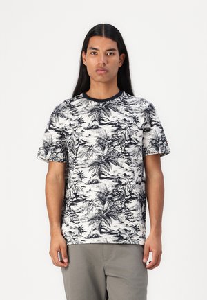 Homme avec de longs cheveux portant un t-shirt à motif tropical noir et blanc et un pantalon gris, se tenant devant un fond blanc uni.