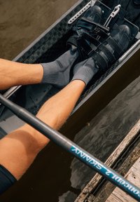 Schwarze Rudersandalen mit Riemen, die über grauen Socken getragen werden, und untergetauchte Beine in einem leichten Boot, sowie ein Ruder gegen eine Holzkante.
