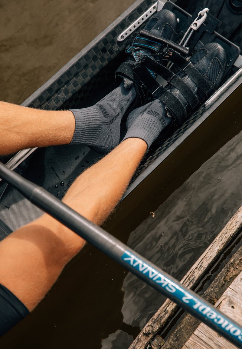 Schwarze Rudersandalen mit Riemen, die über grauen Socken getragen werden, und untergetauchte Beine in einem leichten Boot, sowie ein Ruder gegen eine Holzkante.
