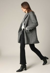 Grijze herringbone wollen jas met gesneden revers, twee voorzakken en een relaxte pasvorm, gecombineerd met aangesloten zwarte broeken en puntige zwarte laarzen.