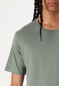 Tricou scurt cu mânecă scurtă, de culoare verde deschis, fabricat din bumbac. Prezintă un gât rotund și o textură netedă, cu cusături vizibile la tiv.