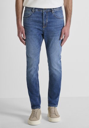 Hombre vestido con vaqueros ajustados azules y zapatillas beige, de pie con las manos relajadas a los lados frente a un fondo liso.