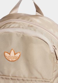 Beige nylon backpack featuring a top handle, front zip pocket, and a brown embroidered logo with three leaves. Smooth texture, durable design.