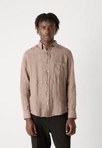 Camicia di lino marrone chiaro con maniche lunghe, colletto a bottone, tasca frontale e sottile texture. Abbinata a pantaloni scuri.