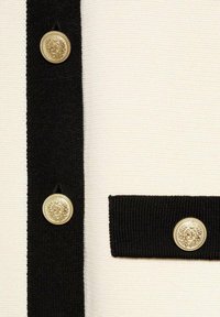 Gros plan d'une bordure noire avec des boutons décoratifs dorés sur un tissu en maille blanche texturée, montrant des détails de rubans verticaux et horizontaux.