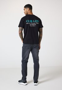 Svart bomull t-skjorte med "CLSR LDN" i Turkis på baksiden, kombinert med grå bukser og svarte joggesko.