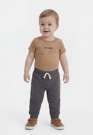 Toddler standing, wearing brown "Best Buddy" shirt, dark gray pants with white drawstring, and brown shoes, smiling slightly.