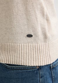 Beige gebreide trui met ribbelzoom en een klein zwart logo. De textuur van de trui is zacht. Draag deze over blauwe denim jeans met contrasterende stiksels.