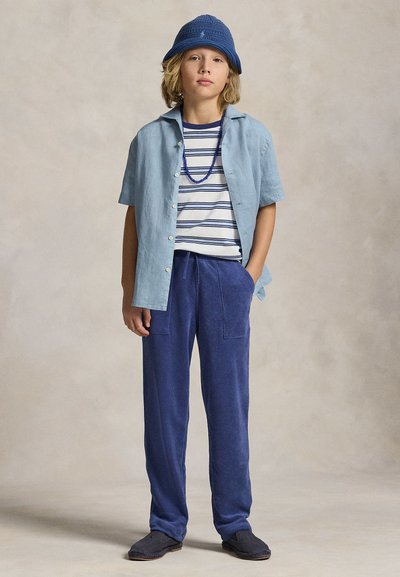 Enfant portant un chapeau seau bleu, une chemise à manches courtes bleu clair sur un t-shirt rayé, un pantalon bleu, des chaussures foncées et un collier de perles bleu.