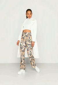 Mulher de pé contra uma parede lisa, vestindo um casaco longo branco, um top cropped branco, calças estampadas multicoloridas e ténis de plataforma brancos.