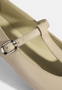 Beige Leder-T-Strap-Schuhe mit matter Oberfläche. Ausgestattet mit einem schmalen Riemen und einer metallischen Schnalle sowie einem leicht abgerundeten Zehendesign.