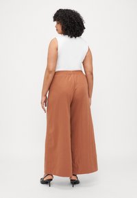 Pantalons larges en tissu brun avec une finition texturée, dotés d'une taille élastique. Portés avec un haut sans manches ajusté blanc.