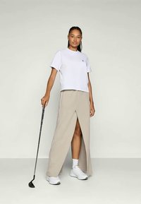 Valge lühikeste varrukatega T-särk, millele on sobitatud beež, lõhestatud maksiseelik. Mudel hoiab golfikeppi, jalas valged jooksujalatsid. Sile kangastekstuur.