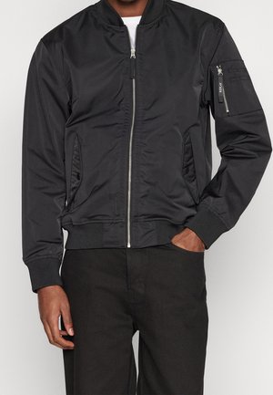 Man wearing black bomber jacket with front zipper, two side pockets, and zippered pocket on left sleeve against gray background.