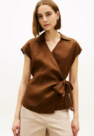 Młoda kobieta z krótkimi brązowymi włosami, ubrana w brązową bluzkę z krótkim rękawem w stylu kopertowym i beżowe spodnie, stoi na tle prostego tła.