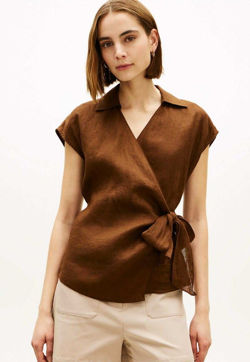 Jeune femme aux cheveux bruns courts portant une blouse marron à manches courtes style portefeuille et un pantalon beige, debout devant un fond uni.