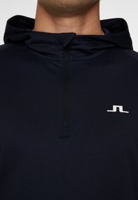 Mörkblå zip-up hoodie i mjukt tyg, med dragkedja fram och en liten vit logotyp på vänster bröst.