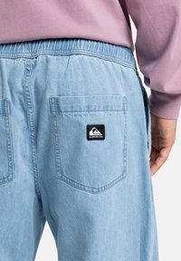 Lyseblå denimbukser med elastisk midje, med to baklommer og et svart logomerke. Glatt tekstur og avslappet passform.