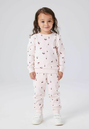 Bambino con capelli lunghi che indossa un coordinato di felpa e pantaloni rosa pallido con stampe di ciliegie, in piedi e sorridente, scarpe bianche.