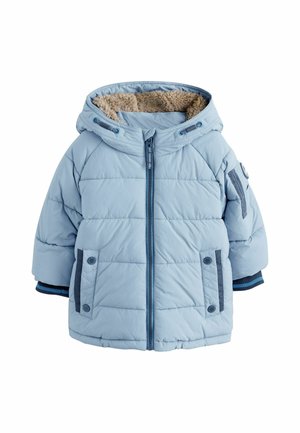 Malé dieťa má na sebe svetlomodrú prešívanú bundu s kapucňou, modrozelené tričko, modré džínsy a tmavomodré tenisky, stojí na jednofarebnom pozadí.