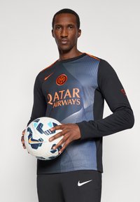 Uomo in maglietta nera e blu dell'Inter che tiene in mano un pallone da calcio Nike bianco e blu su uno sfondo semplice.