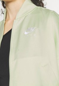 Ljusgrön Nike-jacka med en slät, glänsande textur. Har blixtlåsförslutning och broderad logga med texten "NIKE AIR" i silver.