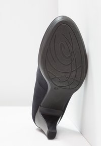 Chaussure à talon haut en cuir suédé noir avec une semelle en caoutchouc noir texturée. Le talon présente une légère courbe et un accent rainuré pour une meilleure adhérence.