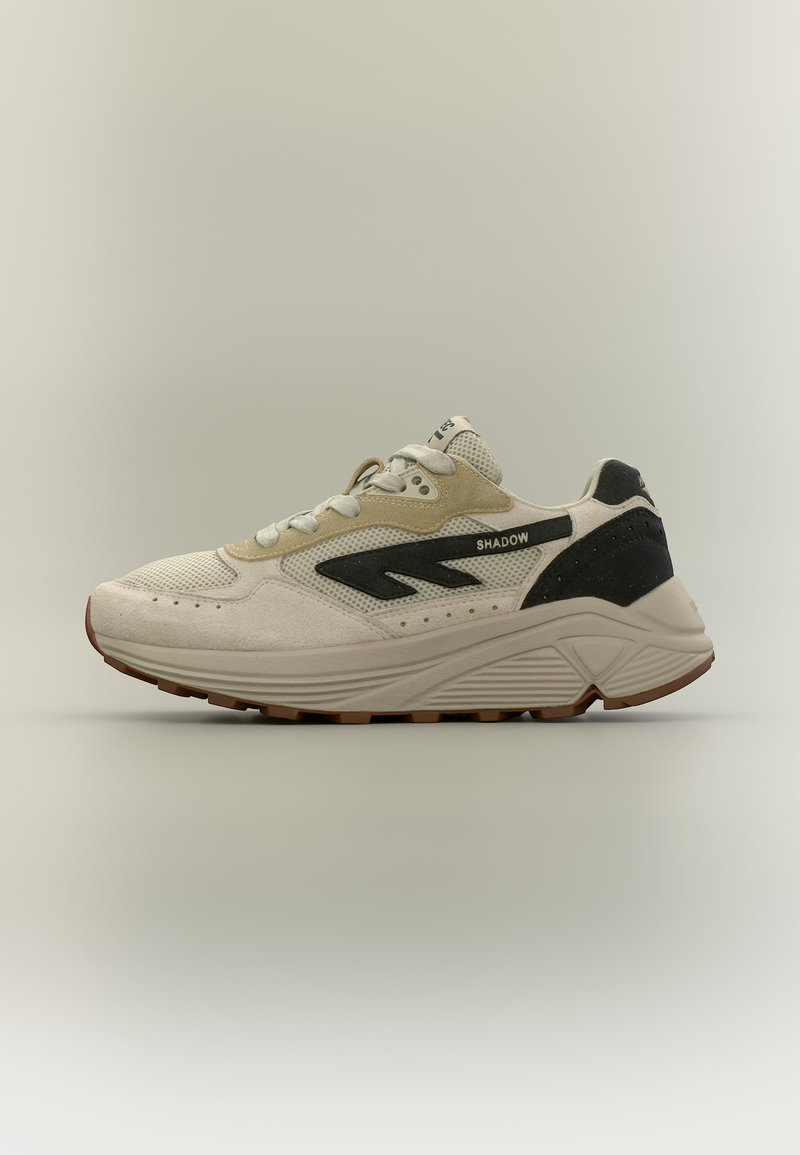 Gaiši bēša un melna sporta apavi ar sieta un zamšādas detaļām, bieza zole un teksts "SHADOW" uz sānu paneļa.