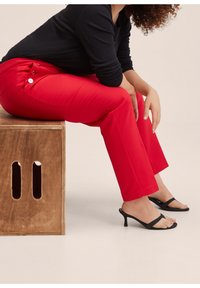 Pantalon rouge sur mesure avec poches latérales et boutons décoratifs, associé à des sandales à talons noires à brides et un haut noir à manches longues.